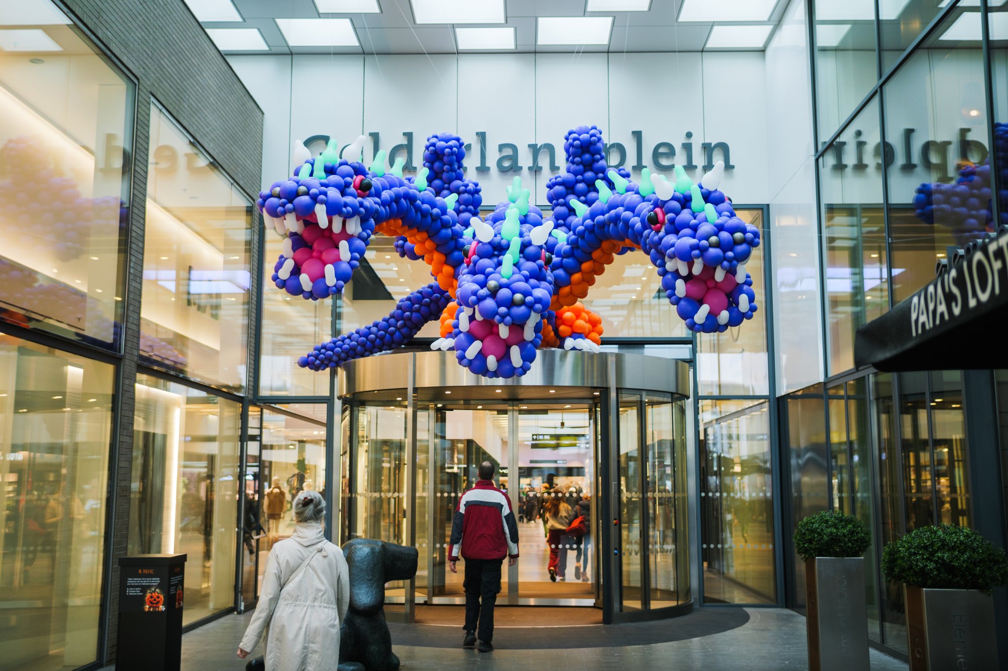 Blue and orange balloon dragon above the revolving doors of Gelderlandplein (2026) — Design: Bidou — Photo: zestien:negen