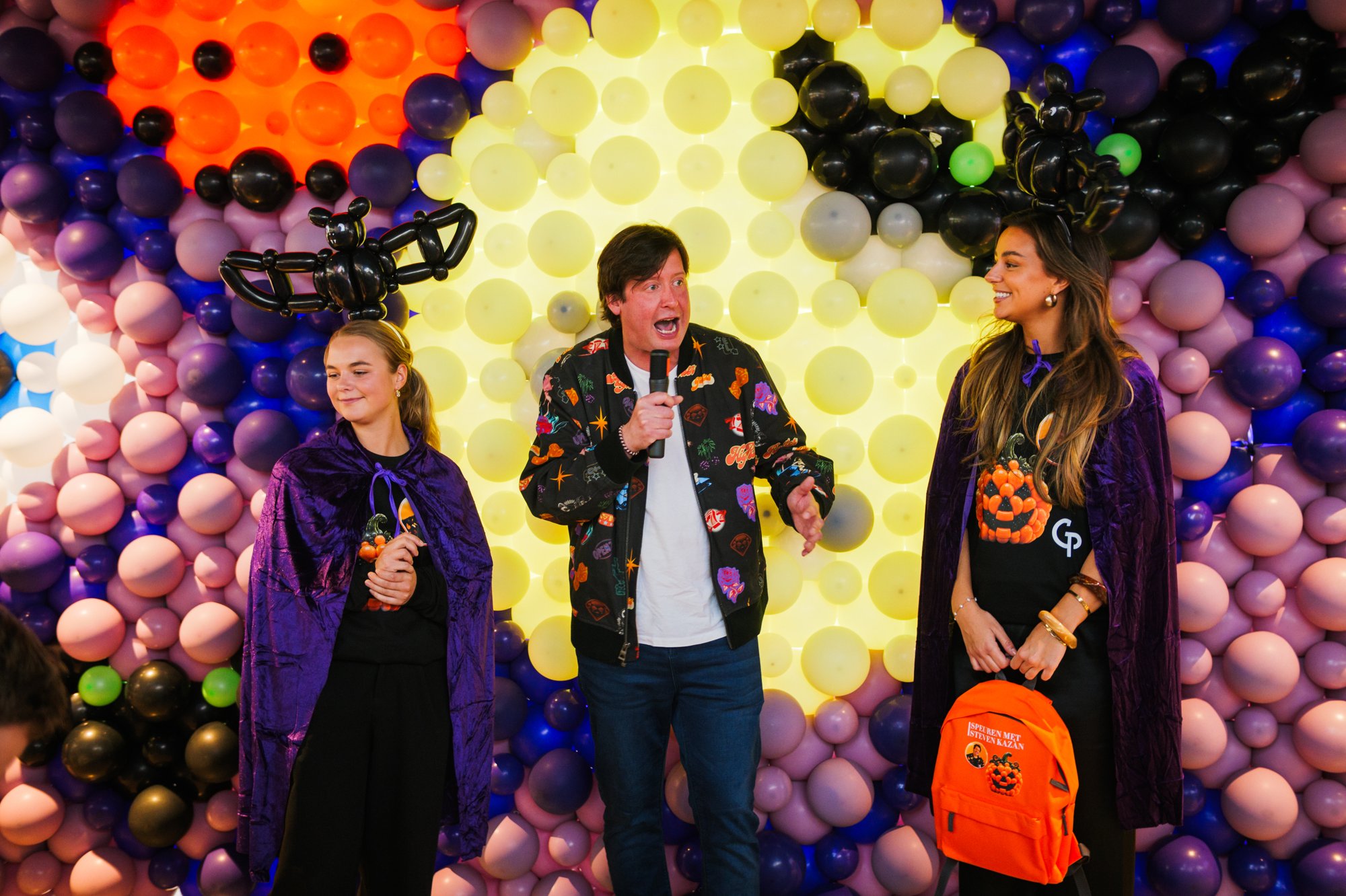 Balloon wall with yellow moon at Gelderlandplein (2026), with host Steven Kazan and hostesses — Design: Micha de Haan and Alex; hostess items Compagnie Les Allumeurs — Photo: zestien:negen